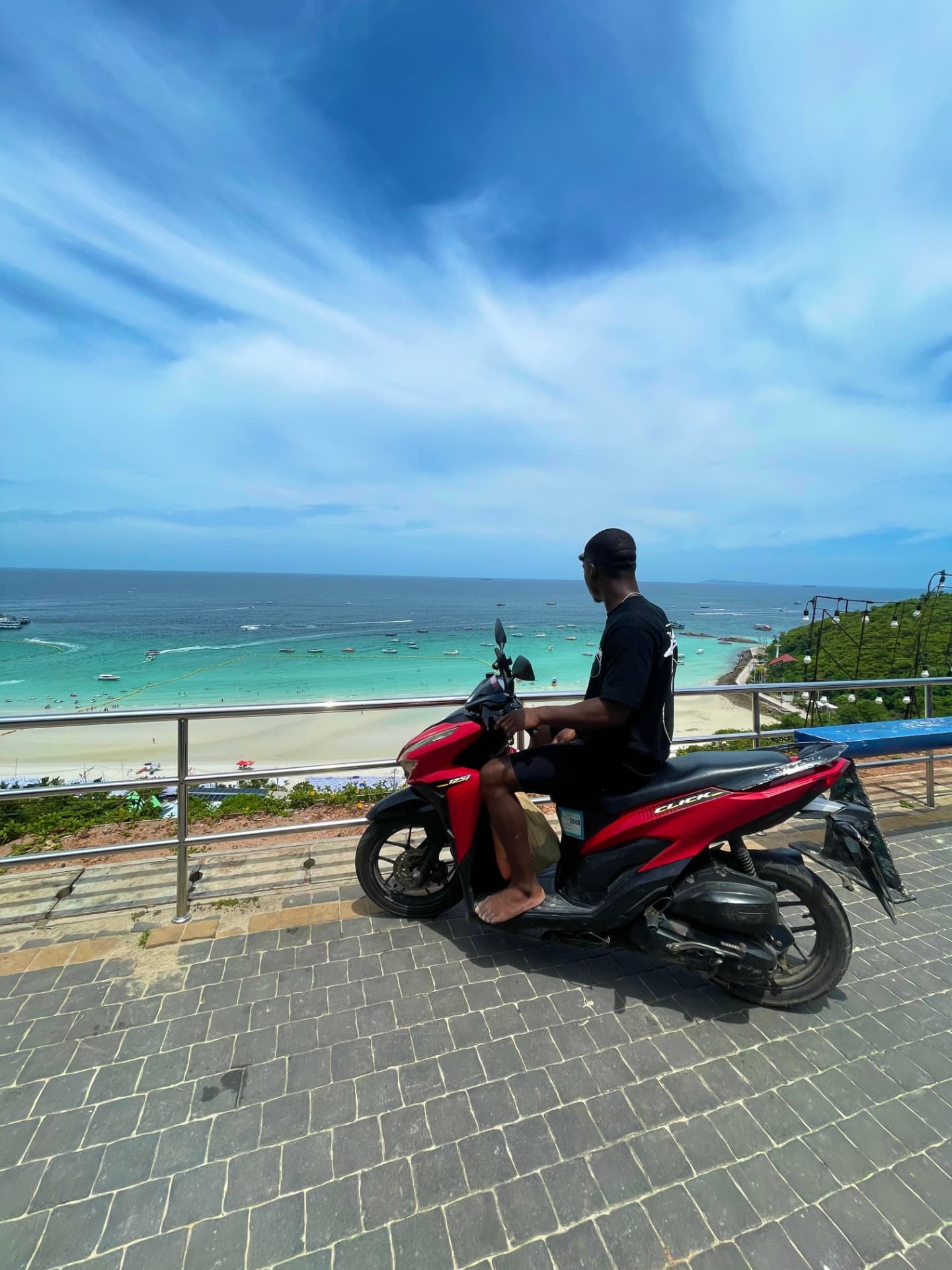 Sitting on a red scooter at a clifftop railing overlooking a turquoise bay and white sand beach